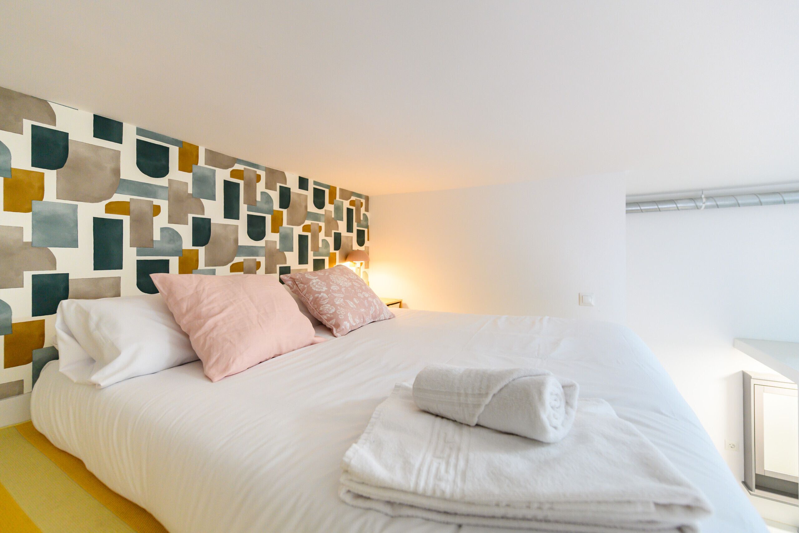 Ett hotellrum med en modern, geometrisk väggdesign, en snyggt bäddad säng med vita sängkläder och ett nattduksbord med en lampa.