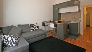 En moderne stue med en grå sofa, et sort gulvtæppe og et køkkenområde med en grå bordplade og hvide skabe.