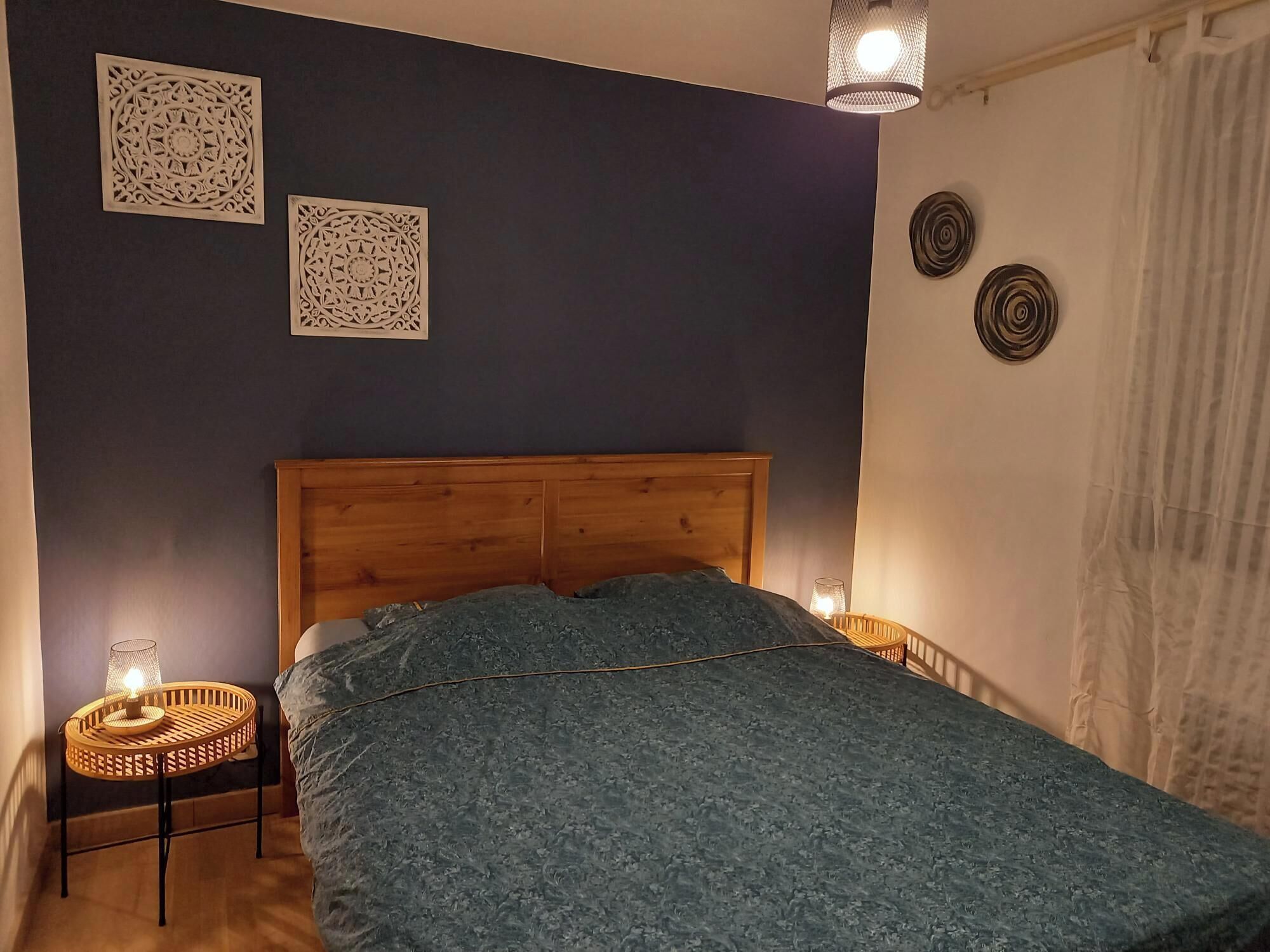 A bedroom with a wooden headboard, a bed with a patterned bedspread, two wall-mounted decorative items, and a small table with a lamp.