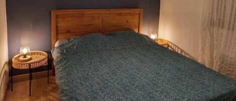 A bedroom with a wooden headboard, a bed with a patterned bedspread, two wall-mounted decorative items, and a small table with a lamp.