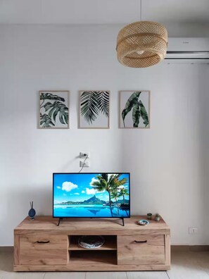 Un moderno salón con un mueble bajo para el televisor de madera, un televisor de pantalla plana mostrando un paisaje tropical, y tres impresiones botánicas enmarcadas en la pared.