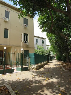 Rua estreita ladeada por edifícios, um portão verde e árvores projetando sombras no chão.
