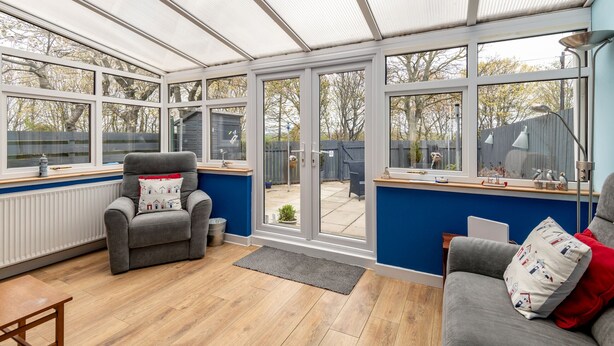 Ein Wintergarten mit Blick auf einen Garten, ein grauer Sessel, ein Holztisch und eine blaue Wand.