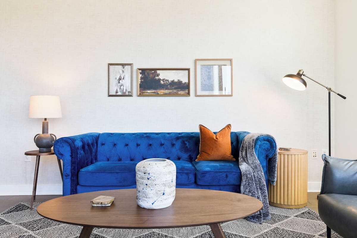 Een woonkamer met een blauwe bank, een ronde salontafel en een lamp. Er hangen drie ingelijste foto’s aan de muur.