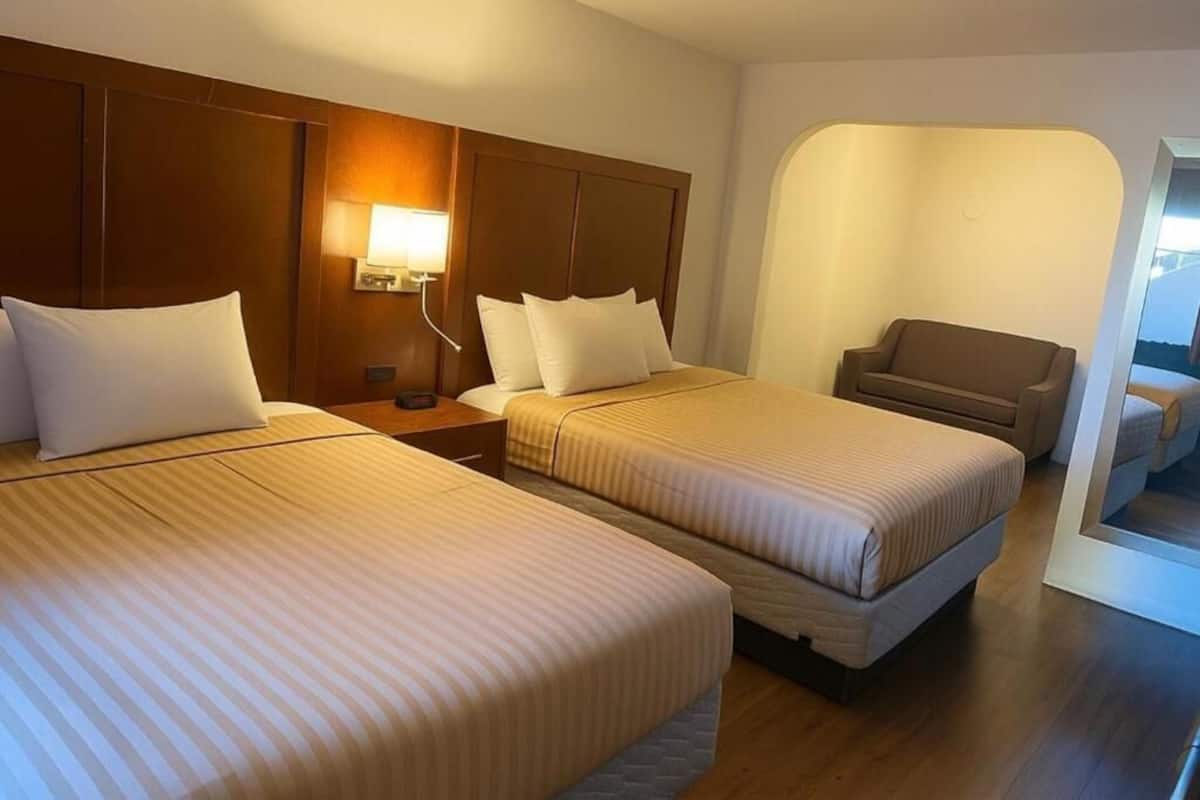 Ein Hotelzimmer mit zwei Betten, einem Nachttisch mit Lampe, einer Sitzecke und Blick durch ein Bogenfenster.