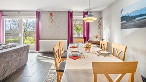 A dining area with a table set for four, chairs, a hanging light fixture, and a view of the outdoors through large windows. - Holiday Home 'Wohnung Wald' with Private Terrace, Garden and Wi-Fi (Ehndorf)