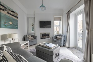 A modern living room with a grey sofa, two armchairs, a coffee table, a flat-screen TV, and a painting on the wall. - Bakers Close | Elie & Earlsferry (Elie)