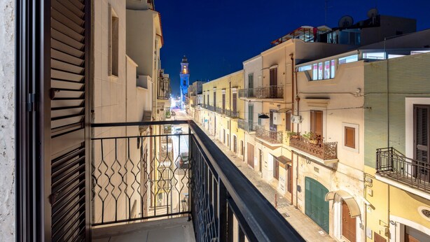 Une rue étroite bordée de bâtiments des deux côtés, un balcon avec une rambarde en métal et, au loin, une tour illuminée.