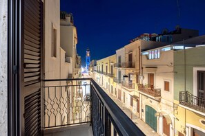 Une rue étroite bordée de bâtiments des deux côtés, un balcon avec une rambarde en métal et, au loin, une tour illuminée.