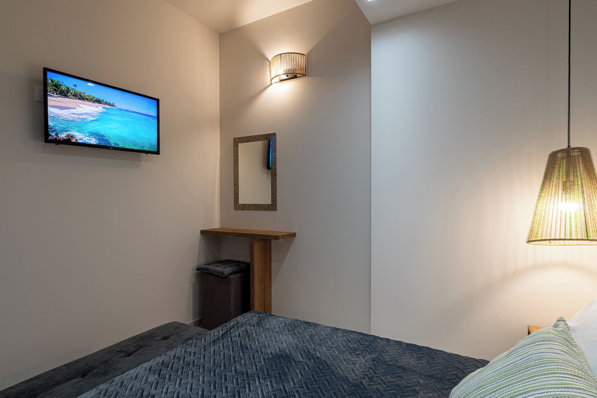 Quarto com cama, televisão na parede, espelho, mesa de madeira pequena e luminária pendente.