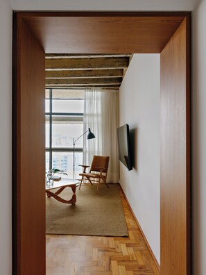 Ein Zimmer mit Holzboden, einem großen Fenster mit Vorhängen, einem Sessel, einem kleinen Tisch und einem Wand-Fernseher.
