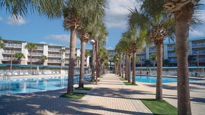 Un resort con un largo paseo junto a la piscina bordeado de palmeras, múltiples piscinas y un edificio de varios pisos con balcones.