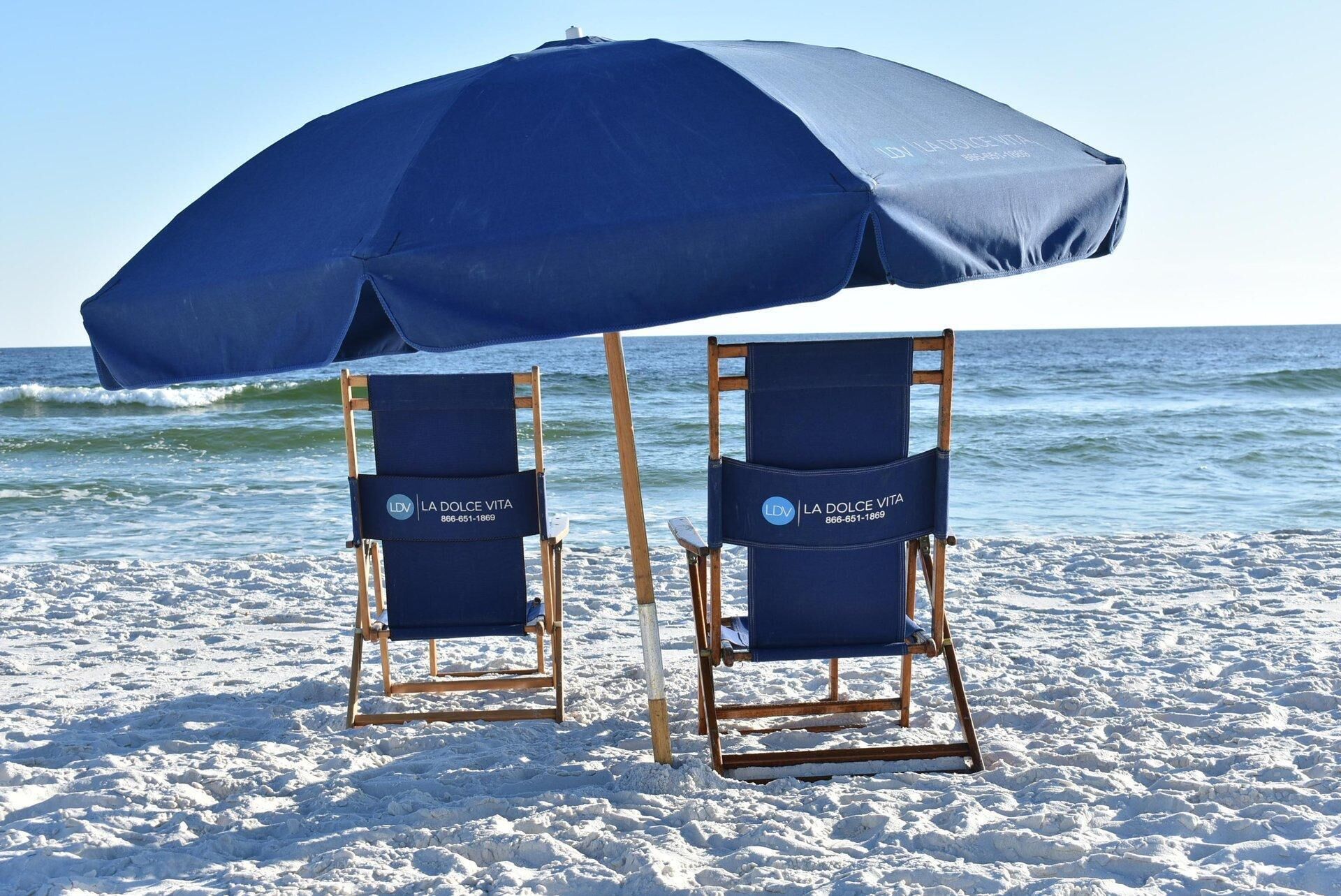 Two beach chairs with a blue umbrella on a sandy beach with the ocean in the background.