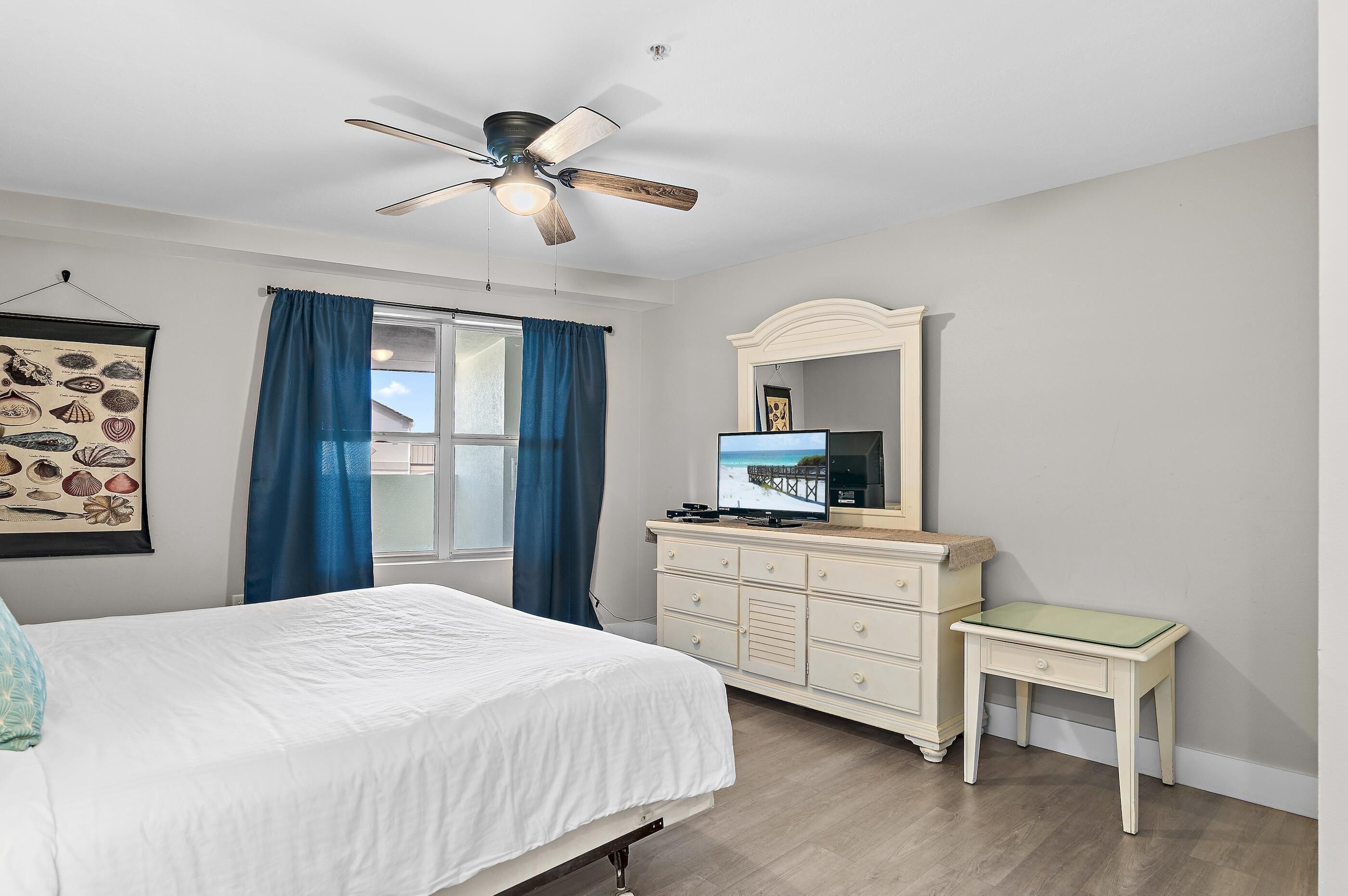 A bedroom with a bed, a dresser, a desk, a ceiling fan, a window with curtains, and a wall-mounted TV.
