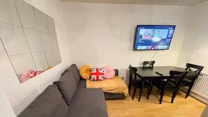 A living room with a grey sofa, a small table with chairs, a flat-screen TV mounted on the wall, and a tiled wall above the sofa. - Brixton Bright Apartment (London)