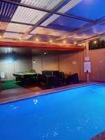 Un área interior con piscina, mesa de billar y sillas, techo con luces y una pared con un cartel.