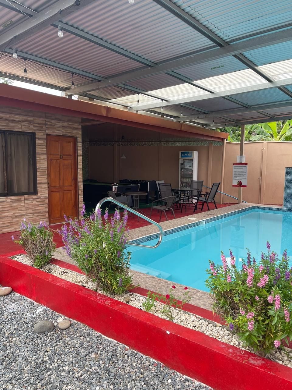 Piscina ao ar livre com borda vermelha, rodeada por plantas e área de estar coberta.