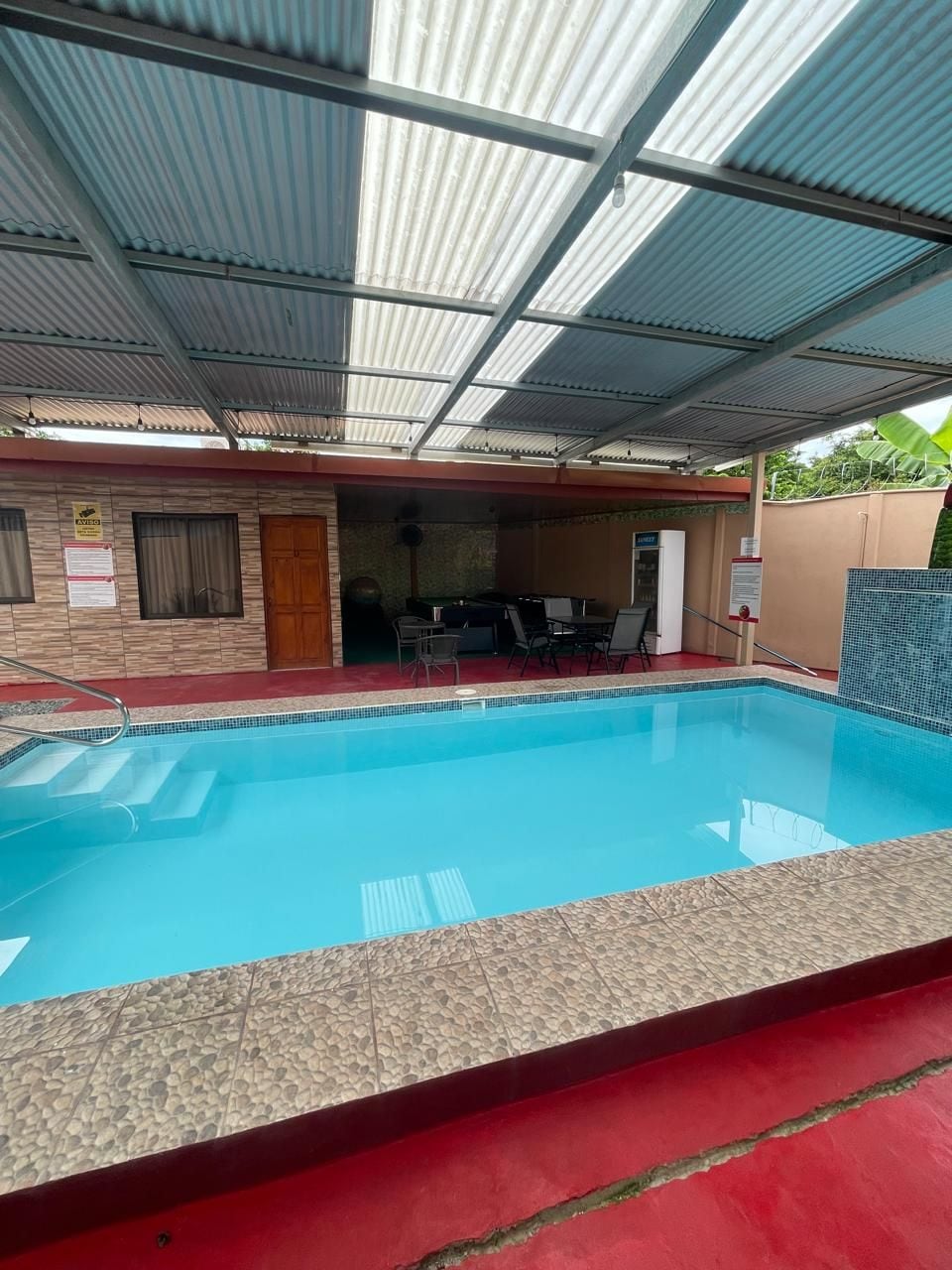 Une piscine avec un bord carrelé, un patio couvert avec des sièges et une porte donnant sur un espace intérieur.