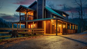 A two-story log cabin with a balcony and a covered porch, surrounded by a wooden fence and a bear sculpture. - Hold My Bear (Pigeon Forge)