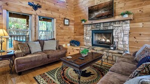A cozy log cabin interior with a stone fireplace, leather sofas, and a large window overlooking greenery. - Hold My Bear (Pigeon Forge)