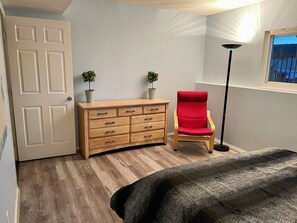 A room with a wooden dresser, a red chair, a bed with a black cover, a floor lamp, and two potted plants. - 3 BR Chantilly Oasis, Pet Friendly, Great Location (Colorado Springs)