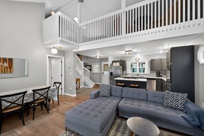 A spacious living area with a grey sectional sofa, a dining table with chairs, a kitchen in the background, and a staircase leading to an upper level. - Peaceful Cable Lake Getaway w/ Dock & Views (Dowagiac)