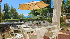 Un área de comedor al aire libre con una mesa preparada para cuatro personas, rodeada de sillas de plástico blancas bajo un paraguas beige.