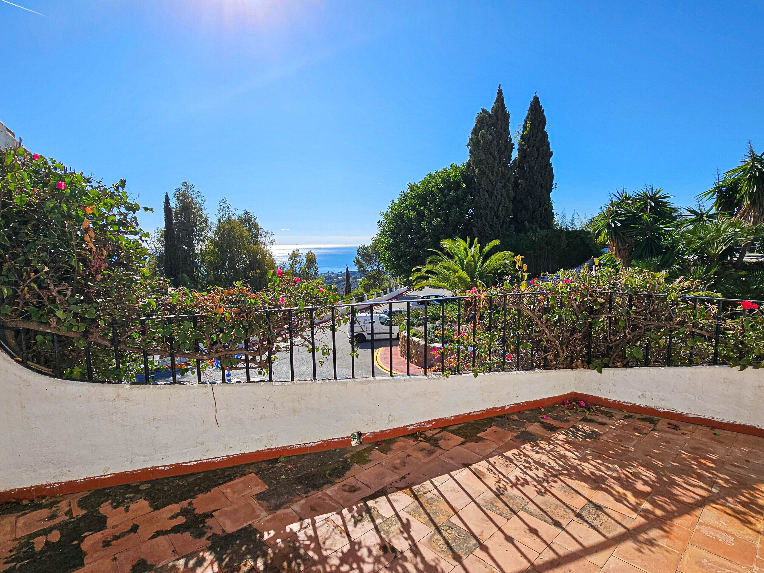 A rooftop terrace with a view of the sea, surrounded by lush greenery and a white railing.