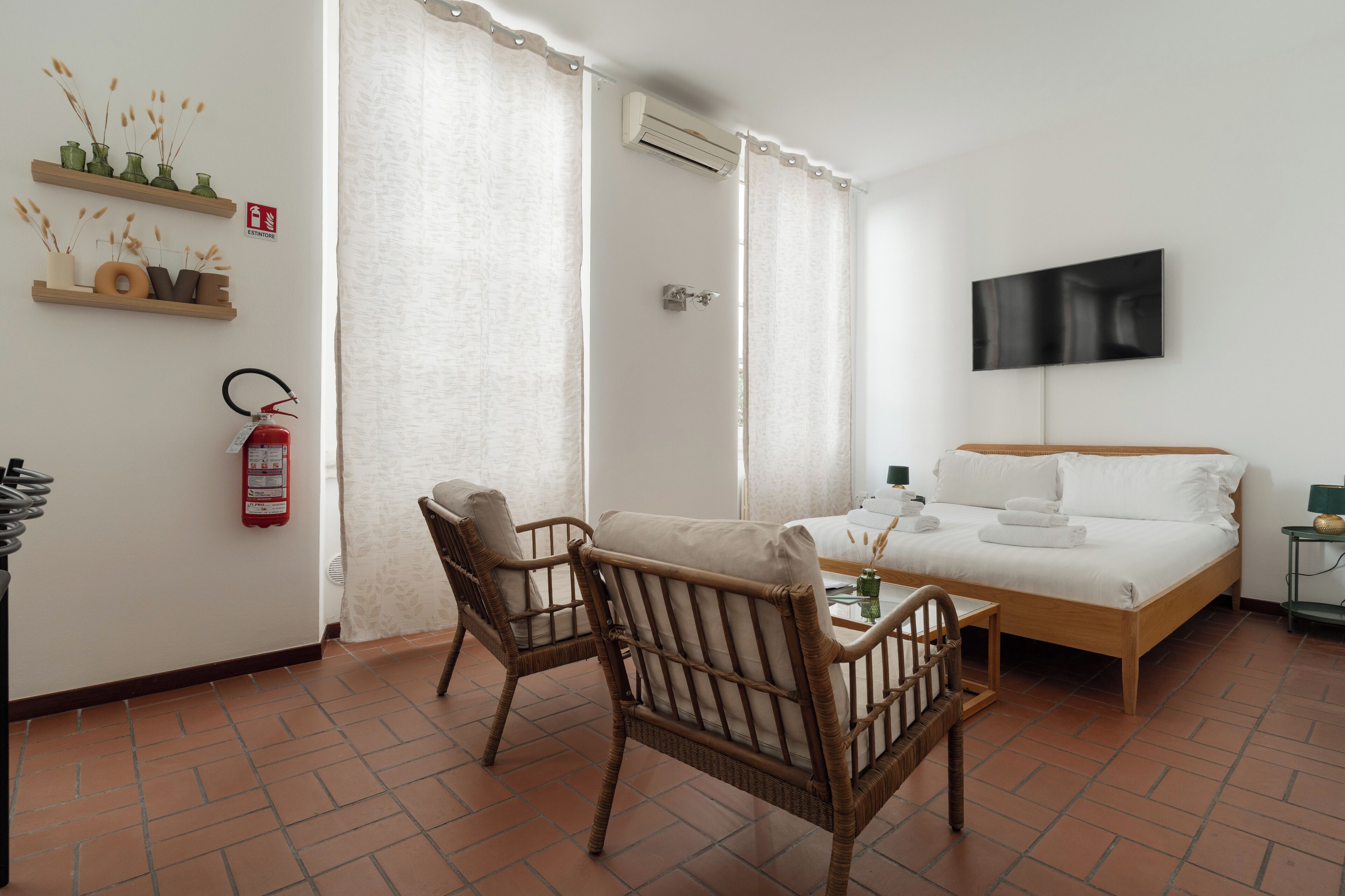 Una camera d'albergo con un letto, due poltrone in vimini, un tavolino, una TV e un estintore appeso al muro.