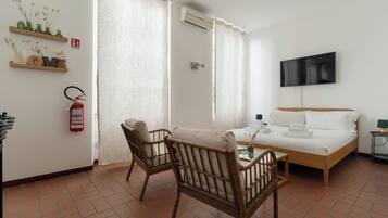 Una camera d'albergo con un letto, due poltrone in vimini, un tavolino, una TV e un estintore appeso al muro.