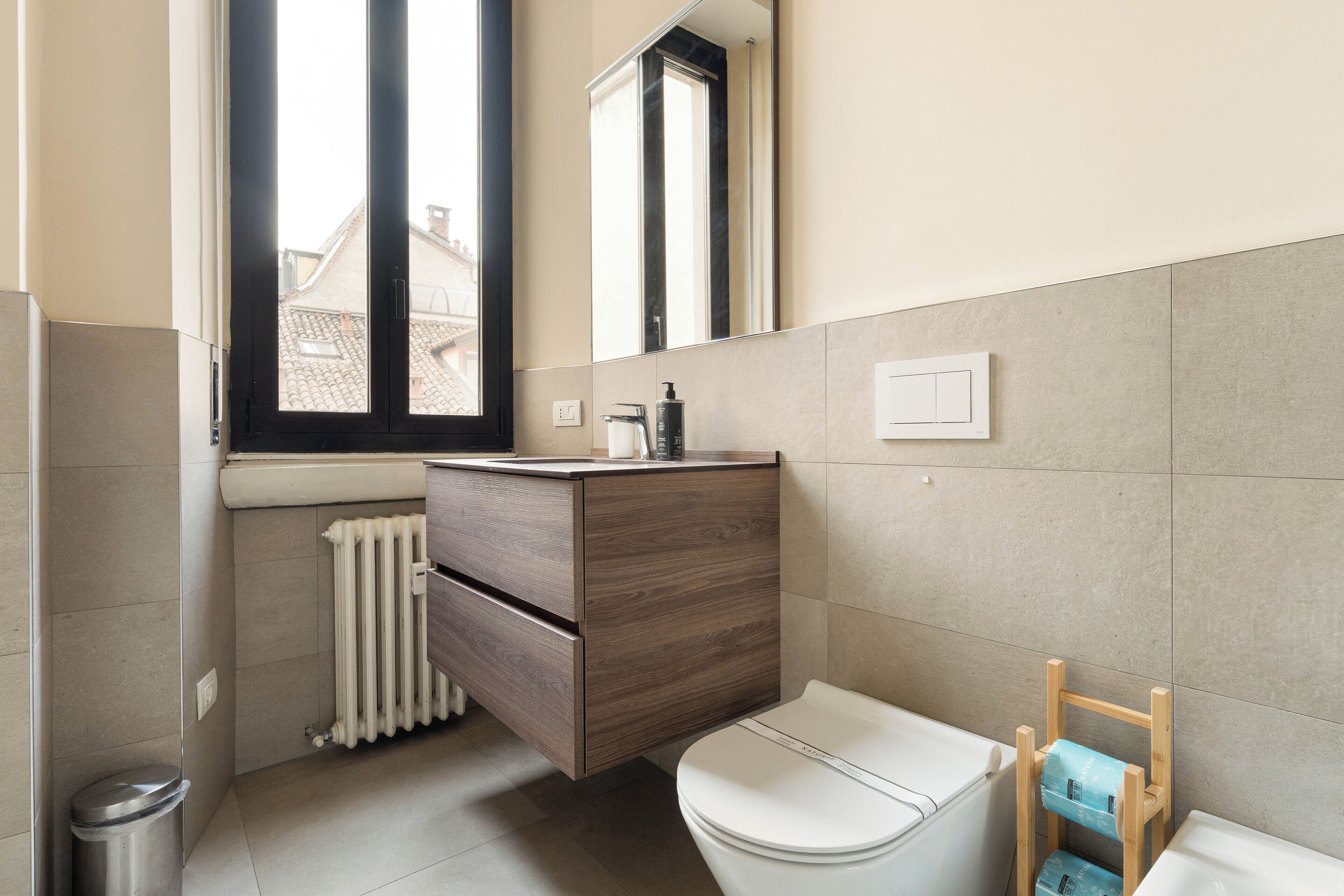 A modern bathroom with a wooden vanity, a toilet, and a window providing natural light.