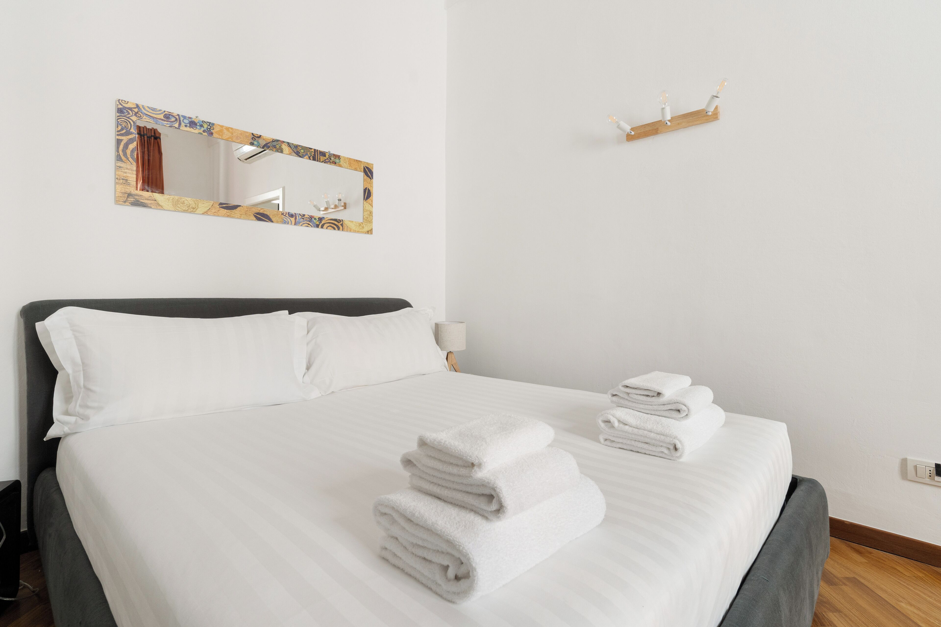 A neatly made bed with white linens and towels, a large mirror with a decorative frame, and a small shelf with figurines.