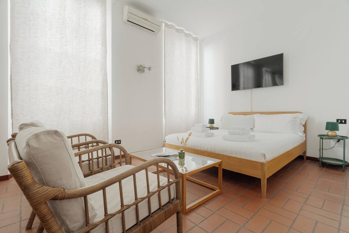 A bedroom with a bed, a chair, a small table, a wall-mounted TV, and a nightstand.