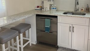 A modern kitchen with white cabinets, a stainless steel microwave, a dishwasher, and a sink with a faucet. There are two bar stools and a decorative rug on the floor.