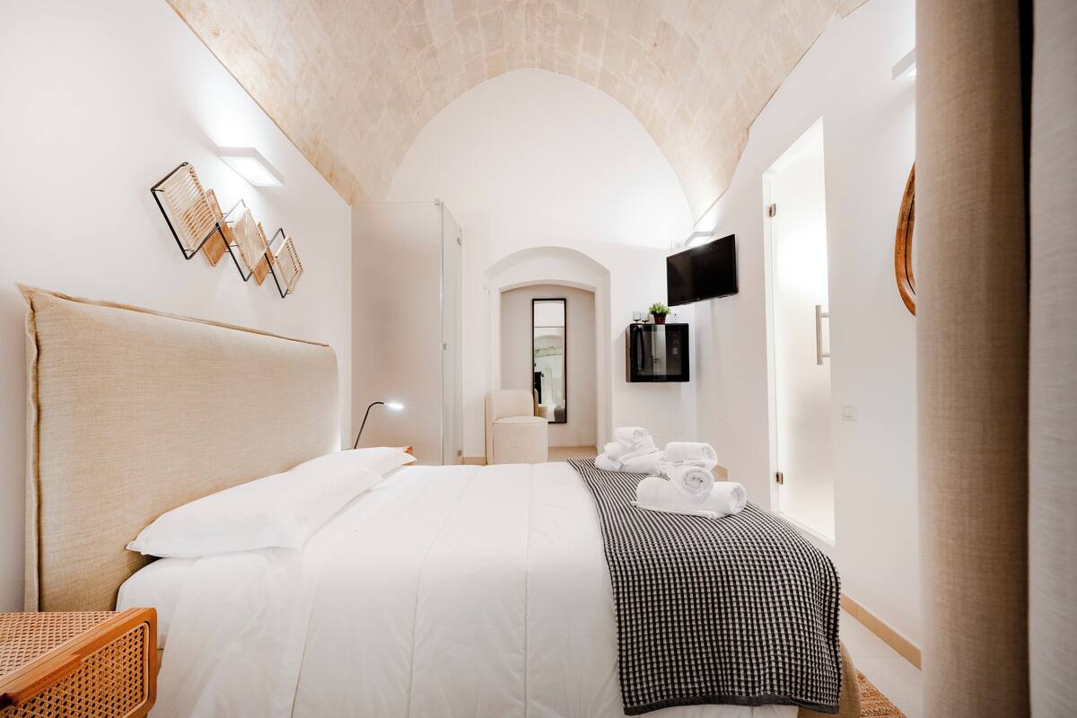 A hotel room with a large bed, a bedside table, a chair, a TV mounted on the wall, and a mirror.