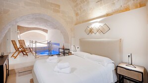 Habitación de hotel con una cama grande, una mesa pequeña con lámpara, una silla, un escritorio y vistas a una piscina a través de un arco.
