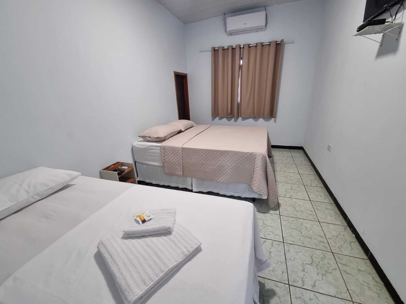 A small, clean room with two beds, a window with curtains, and a wall-mounted air conditioner.