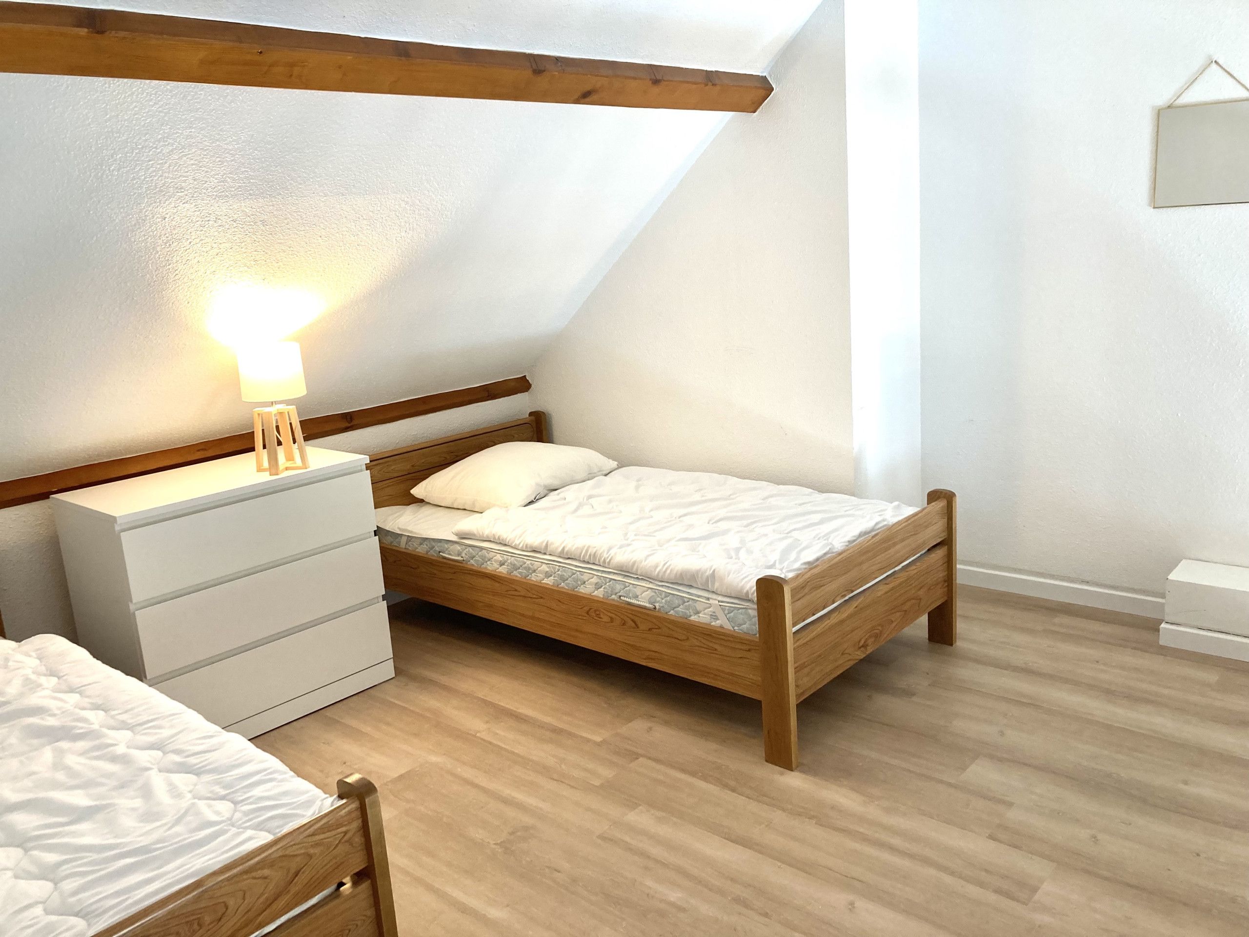 A bedroom with a sloped ceiling, a wooden bed frame, a bedside table with a lamp, and a white dresser.