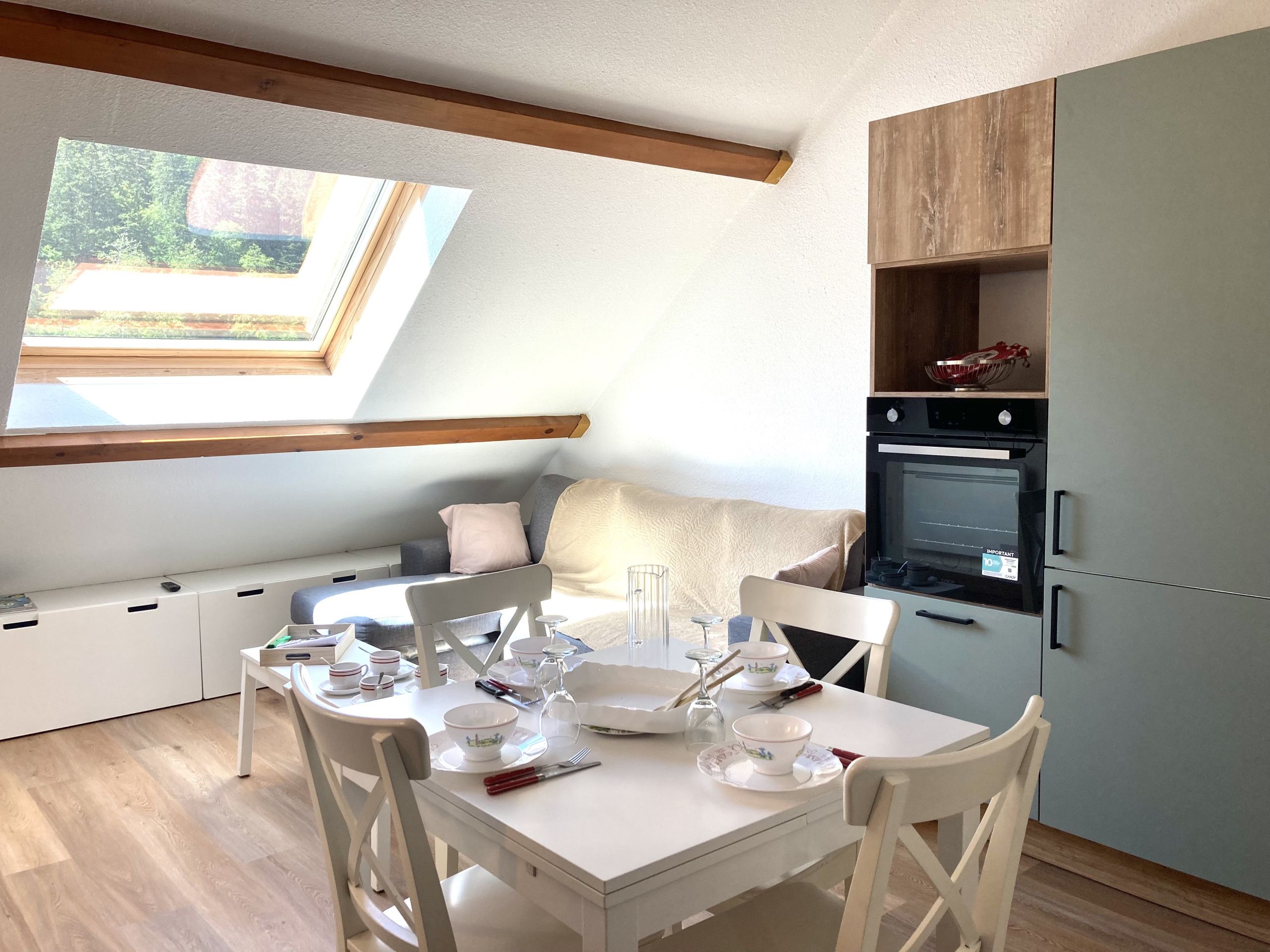 Un coin repas avec une table dressée pour deux personnes, un four encastré et une fenêtre donnant sur un paysage verdoyant.