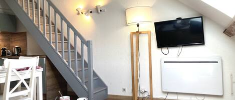 Un espace de vie moderne comprenant un escalier, un coin repas avec des meubles blancs, un téléviseur à écran plat fixé au mur et un lampadaire.