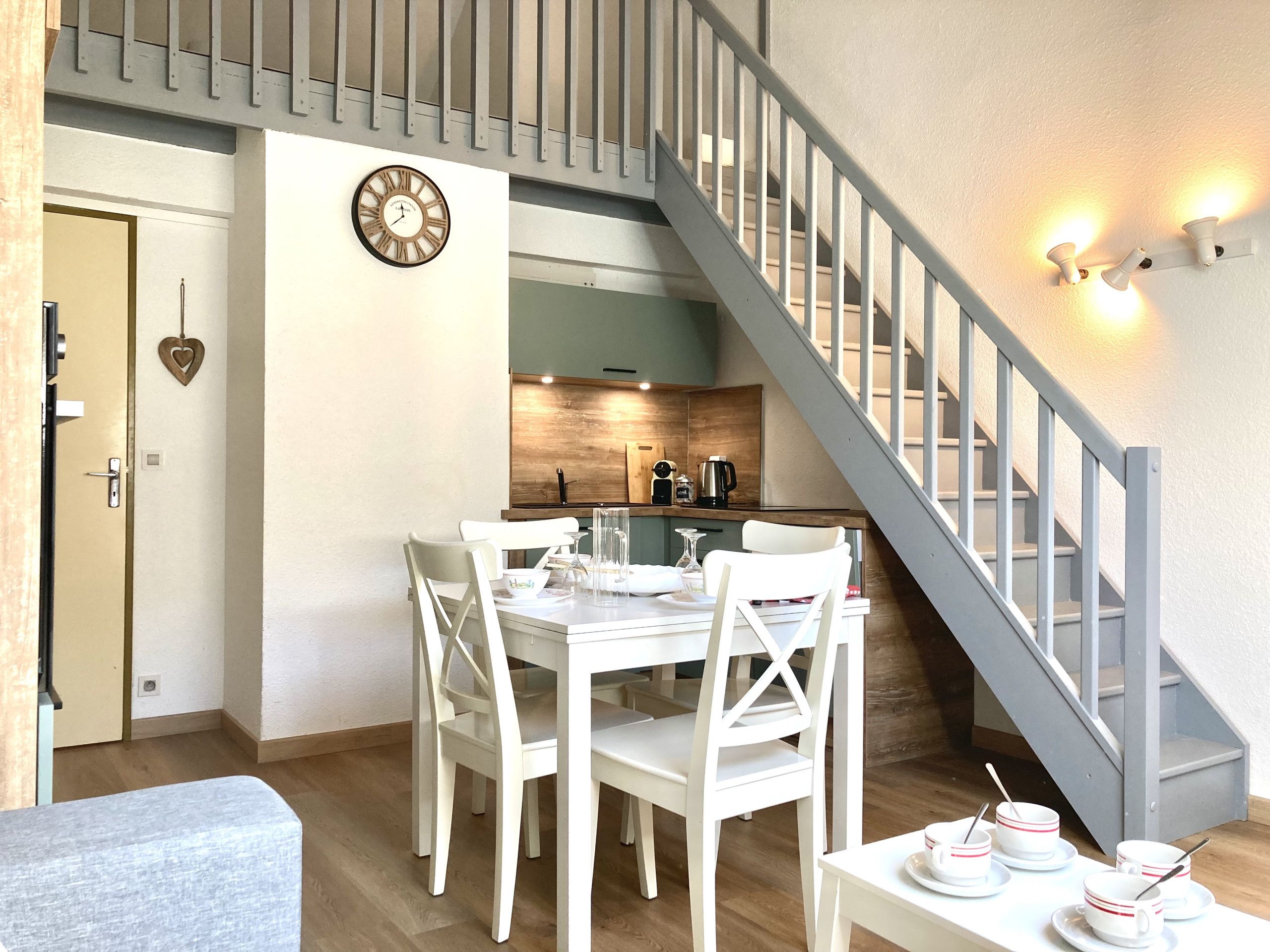 A dining area with a white table and chairs, a kitchen with a wooden countertop, and a staircase with a gray railing.