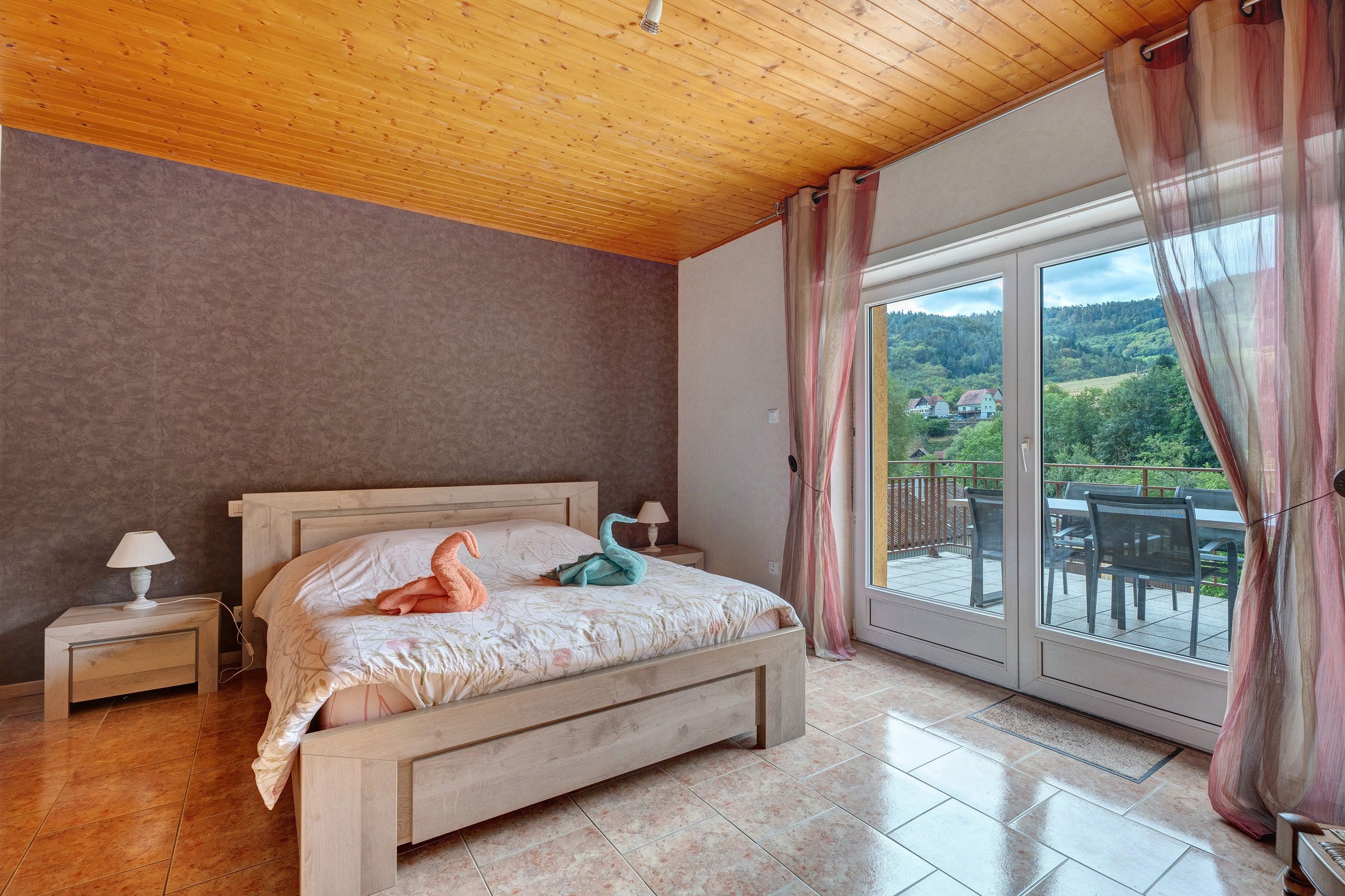 Un dormitorio con techo de madera, una cama con colcha blanca, dos mesitas de noche con lámparas y vistas a un balcón con mobiliario exterior.