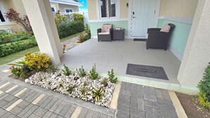 Ein überdachter Außenbereich mit zwei Sitzbereichen, einer Tür und einem kleinen Garten mit Pflanzen und einem Weg.
