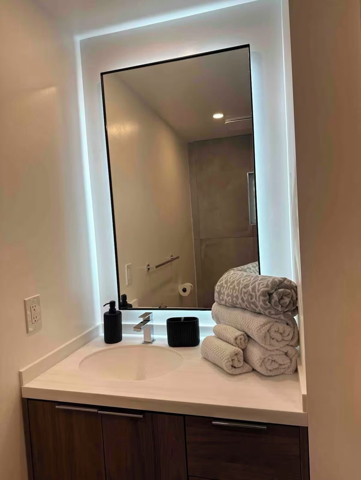 A modern bathroom with a large mirror, white countertop, and a stack of towels.