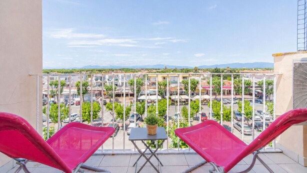 Ein Balkon mit zwei roten Stühlen und einem kleinen Tisch, mit Blick auf einen Parkplatz und Gebäude.