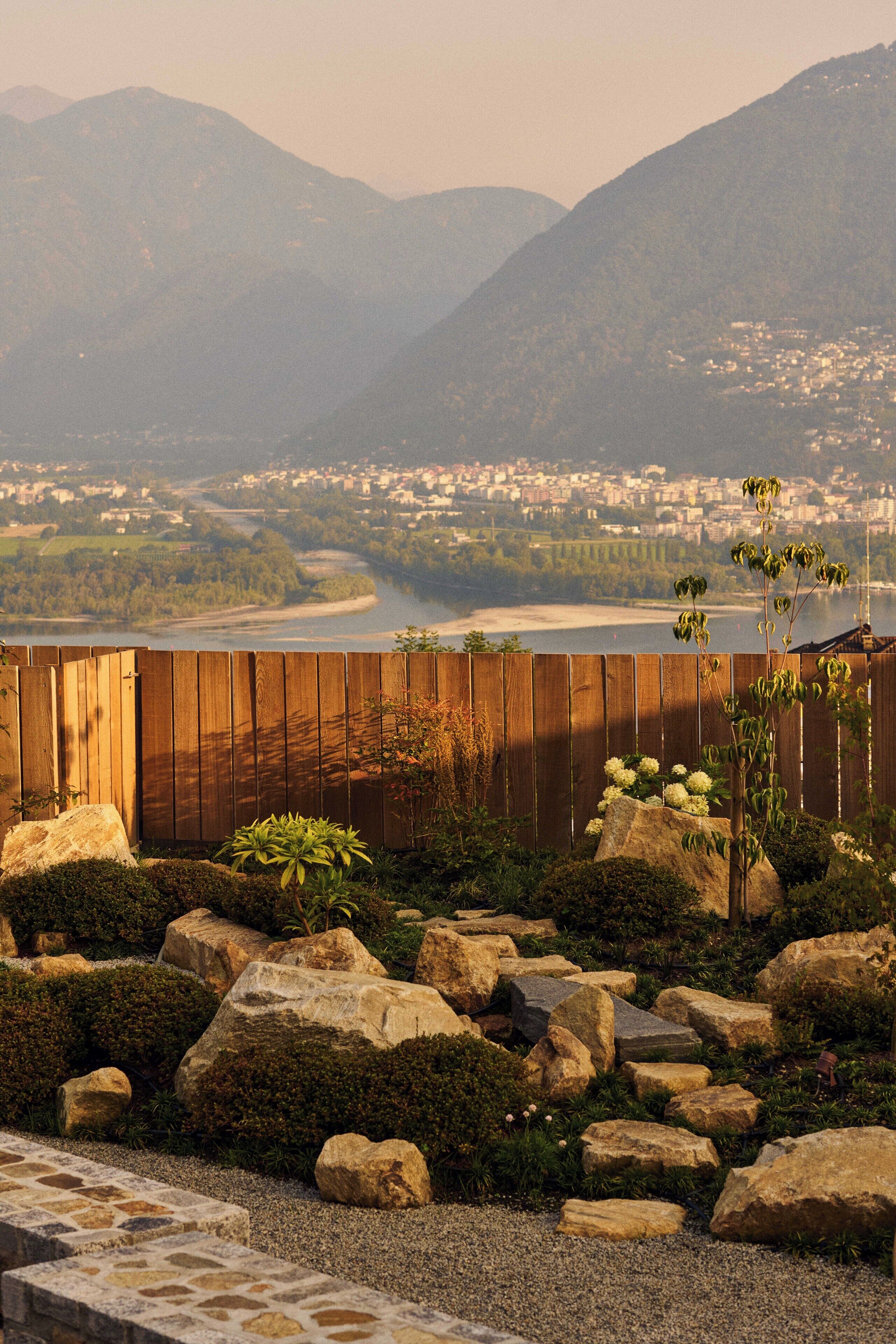 A view of a mountainous landscape with a wooden fence in the foreground, a river in the middle ground, and a town in the background.