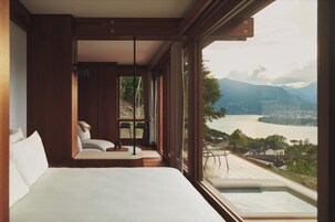 Un dormitorio con una cama grande, una zona de estar con una silla y una mesa, y vistas a un lago y montañas a través de amplios ventanales.