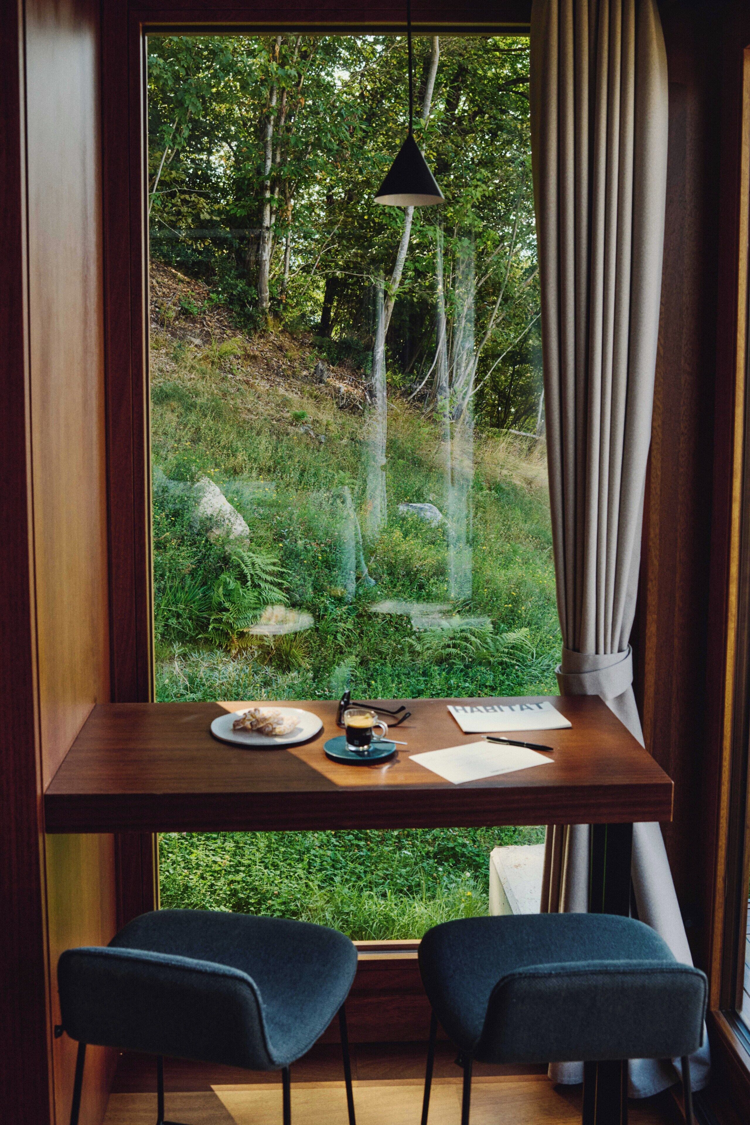 A room with a large window overlooking a forested area, a wooden desk with two chairs, and a hanging lamp.