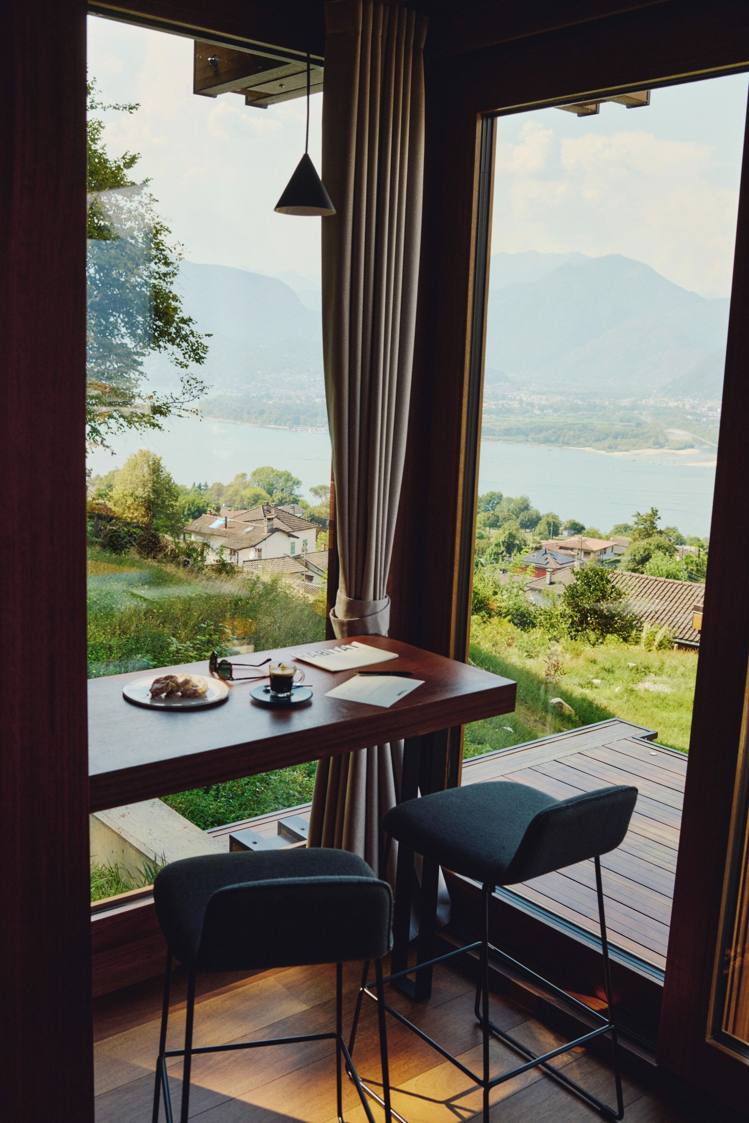 A room with a wooden table, two chairs, a view of a lake and mountains, and a hanging light fixture.