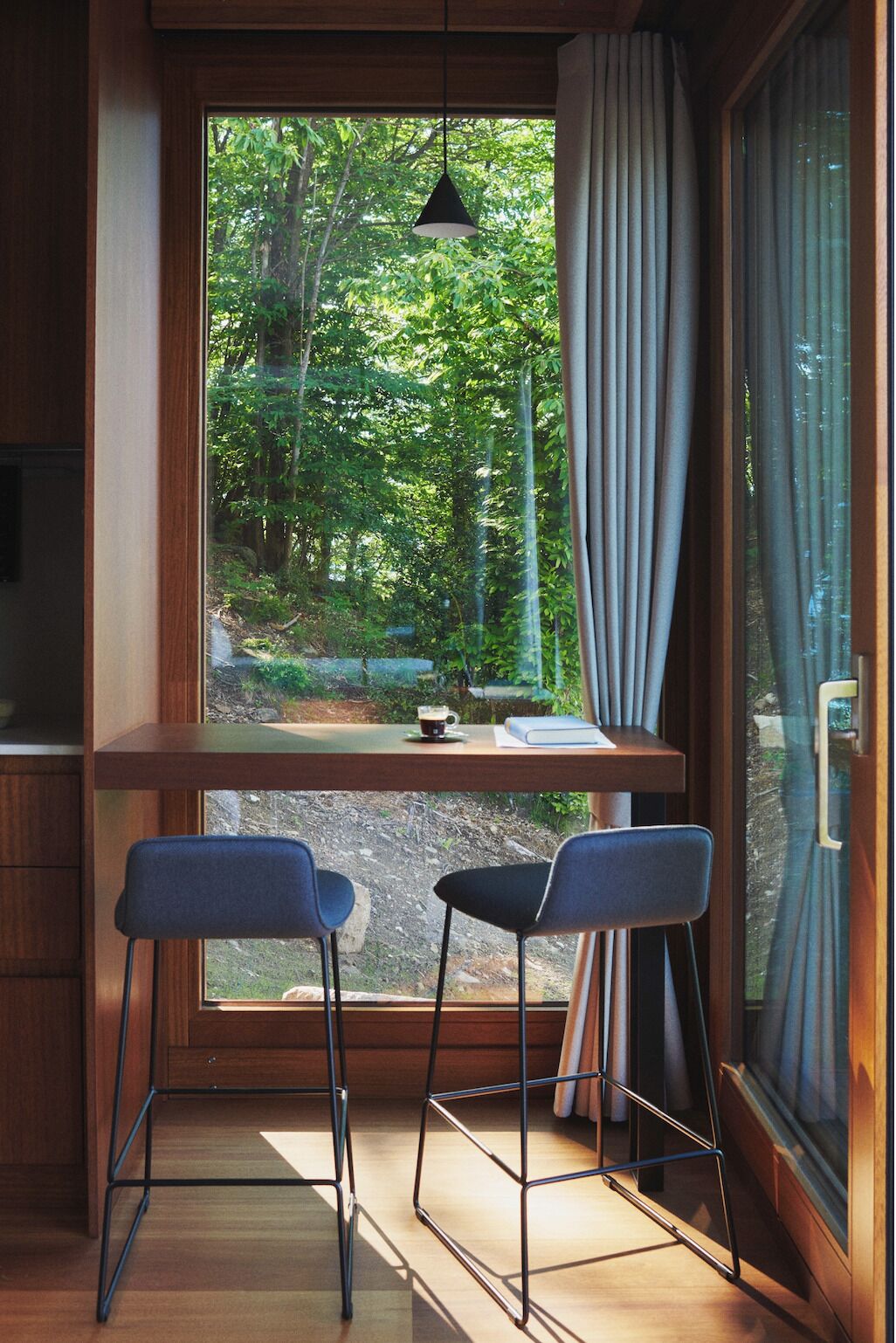 A room with a large window, two blue bar stools, a wooden table with a book and a cup, and a hanging pendant light.